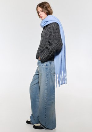 Maglione grigio texture, sciarpa oversize azzurra, jeans denim a gamba larga, scarpe in bianco e nero, posa in piedi, sfondo semplice.