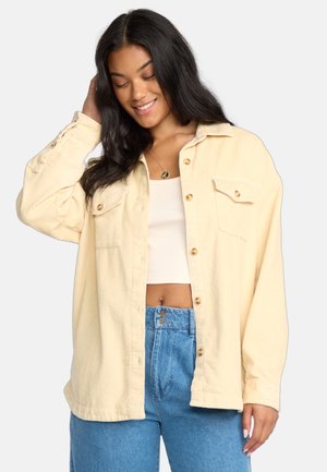 Veste en velours côtelé jaune clair avec un col, deux poches poitrine et des boutons dorés, associée à un crop top blanc et un jean taille haute bleu.
