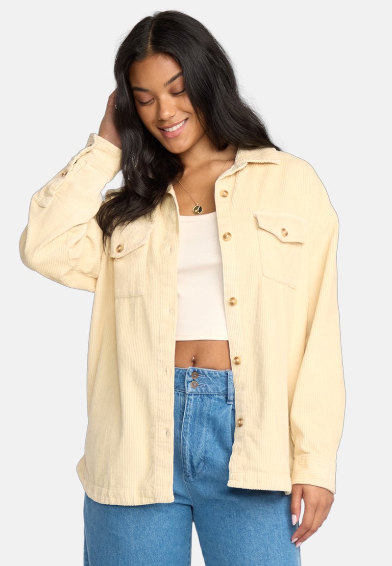 Giacca camicia in corduroy giallo chiaro con colletto, due tasche sul petto e bottoni dorati, abbinata a un crop top bianco e jeans a vita alta blue.