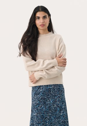 Beige knitted sweater with ribbed cuffs and hem, paired with a long, patterned navy skirt featuring blue and white accents.