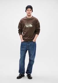 Brun sweatshirt med grafisk tryck, blå denimjeans och svarta skor. Modellen bär en svart stickad mössa och står mot en vit bakgrund.