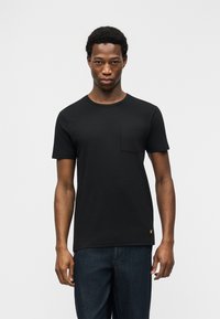 Νέος άνδρας που φοράει απλό μαύρο κοντομάνικο μπλουζάκι με τσέπη στο στήθος και σκούρο μπλε τζιν, όρθιος μπροστά σε λευκό φόντο.