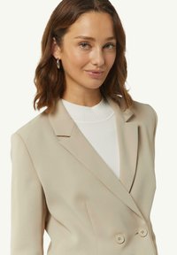 Beige blazer met een dubbelgeknoopt ontwerp, met gelapte revers en twee knopen aan de voorkant, gedragen over een wit geribbeld sweater. Stroeve stoftextuur.