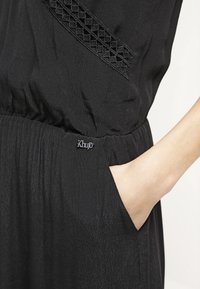 Gros plan d’une personne portant une robe noire avec une ceinture texturée et un détail en dentelle, main placée dans une poche latérale.