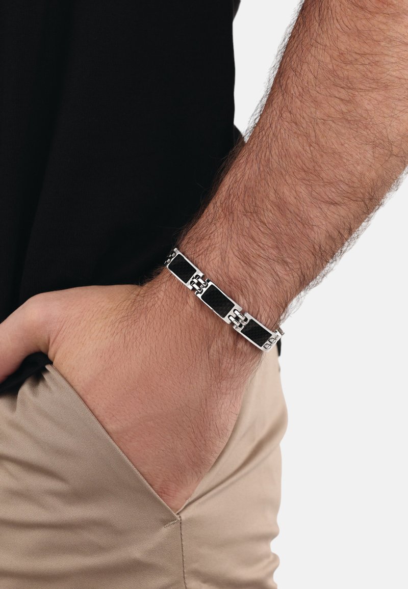 Pulsera de cadena negra y plateada con eslabones rectangulares, detalles texturizados en negro y un acabado pulido, llevada en una muñeca.