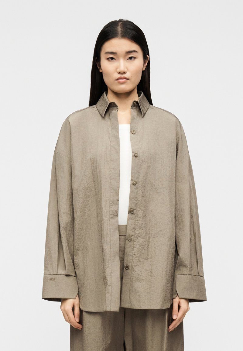 Νεαρή γυναίκα που φοράει υπερμεγέθη πουκάμισο σ ε χρώμα ταυ με φαρδιά μανίκια πάνω από λευκό τοπ και ταιριαστό παντελόνι ταυ, στέκεται μπροστά σε απλό φόντο.