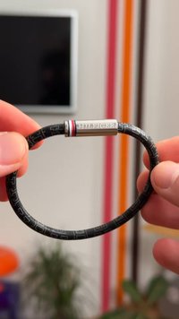 Μαύρο ελαστικό βραχιόλι με ανάγλυφο σχέδιο, μεταλλική πόρπη με το λογότυπο "TH", κόκκινες και μπλε λεπτομέρειες. Λείος υφή, ευέλικτο σχέδιο.