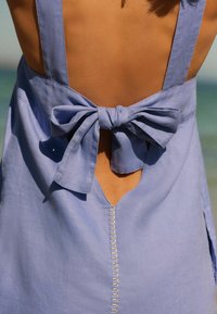 Hellblaues Leinenkleid mit einem Schlüsselrückenausschnitt, verziert mit einer Schleife und dekorativen Nähten, die durch die Mitte verlaufen. Weicher Stoff.