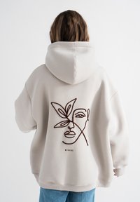 Beige übergroßer Hoodie mit Kapuze und Kordelzug. Auf der Rückseite befindet sich ein dunkelschokoladenbraunes gesticktes Design eines Gesichts und eines Blattes. Mischgewebe aus Baumwolle.