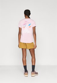 Ροζ ανοιχτό t-shirt με κοντά μανίκια, που απεικονίζει ένα γραφικό βουνό σε λευκό, μπλε και πορτοκαλί χρώμα. Συνδυάζεται με κίτρινα σορτς και παπούτσια πεζοπορίας.