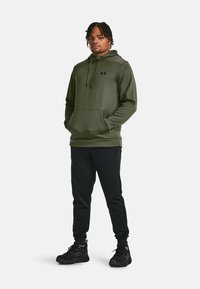 Olivengrøn hættetrøje med kængurulomme og snoretræk i hætten, parret med sorte tapered joggers og sorte sportsko.