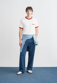 Λευκό T-shirt με κόκκινη επένδυση και λογότυπο, ανοιχτό μπλε πουλόβερ δεμένο γύρω από τη μέση, σκούρο μπλε αθλητικό παντελόνι και λευκά αθλητικά παπούτσια με ταμπά σόλα.