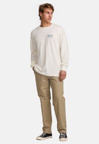 Long-sleeve cream shirt with blue logo on chest, paired with beige pants and black-and-white sneakers. Simple design and relaxed fit.