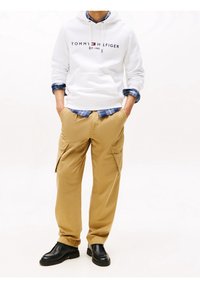 Felpa bianca con logo "TOMMY HILFIGER", camicia a quadri blu sotto, pantaloni cargo beige e scarpe nere con suola a texture.