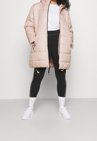 Person klädd i en ljusrosa puffjacka över en vit skjorta, svarta Nike leggings, vita strumpor och vita sneakers, står med benen isär.