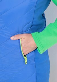 Blå isolerad jacka med diagonala sömmar, en grön detalj runt kragen och en sidoficka med dragkedja i en stark färg.