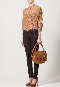 Bluzka z długim rękawem w pomarańczowy kwiatowy wzór, zestawiona z ciemnymi dopasowanymi dżinsami i brązową skórzaną torebką. Modelka ma na sobie brązowe wysokie obcasy.