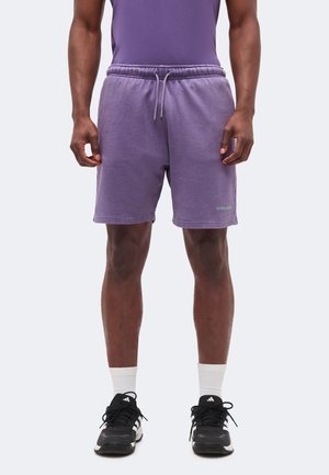 Άνδρας με μοβ σορτς, μαύρα αθλητικά παπούτσια Adidas και λευκές κάλτσες μέχρι τον αστράγαλο, στέκεται με τα χέρια χαλαρά στα πλάγια σε μονόχρωμο φόντο.