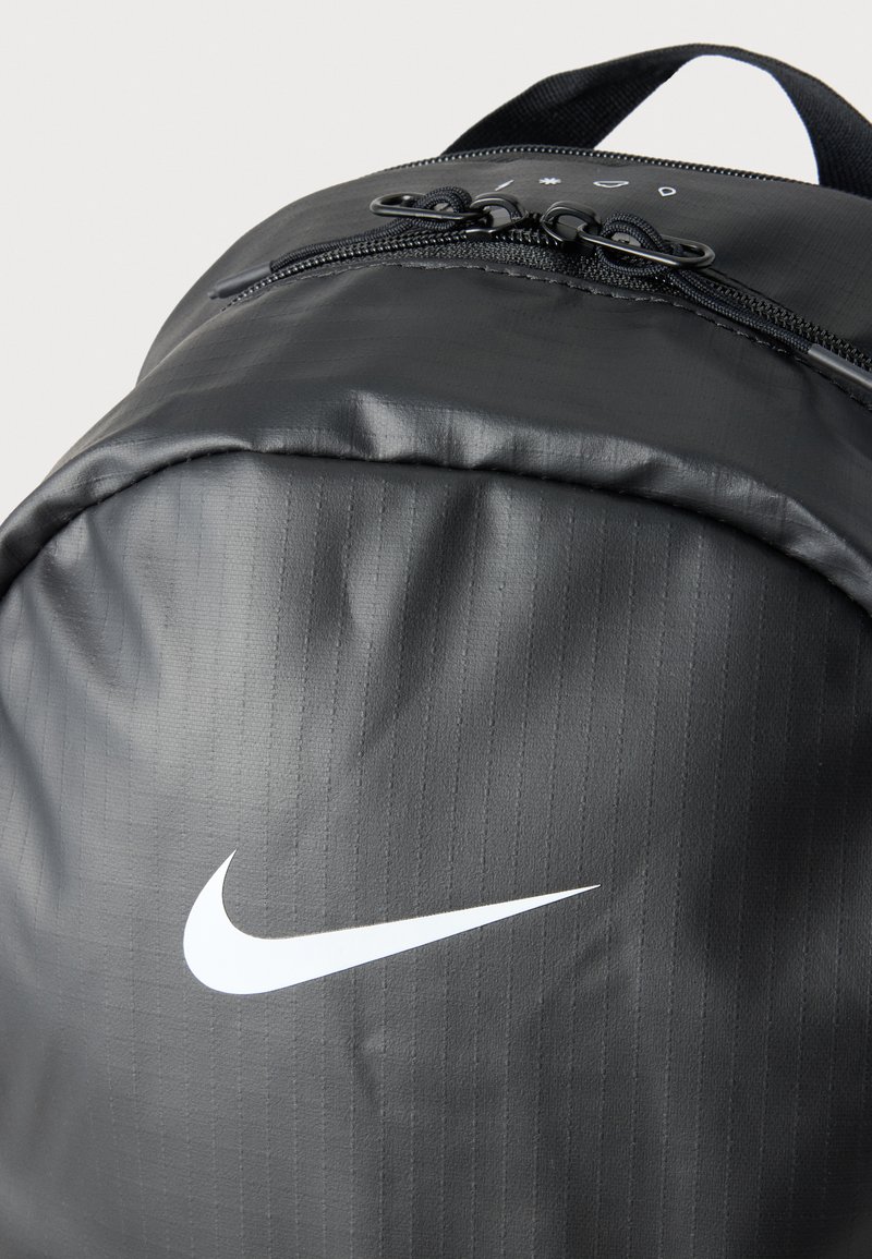 Close-up of a black Nike backpack with dual zipper pulls and a white Nike swoosh logo on a textured fabric surface.