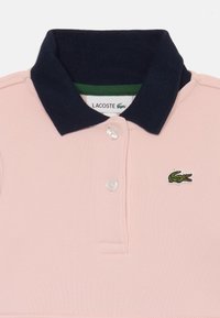 Różowa koszulka polo z ciemnoniebieskim kołnierzykiem, dwoma białymi guzikami i zielonym haftowanym logo krokodyla Lacoste na piersi.