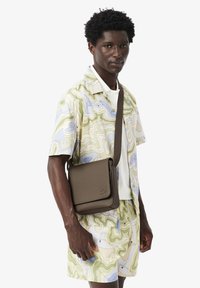 Uomo che indossa una camicia e pantaloncini con stampa topografica pastello, con una borsa a tracolla marrone con patta e logo in rilievo.