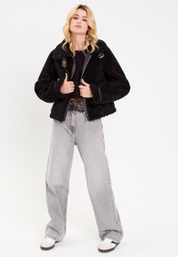 Schwarze Kunstfelljacke mit kurzem Schnitt, weiten Ärmeln und Druckknopf. Kombiniert mit hellgrauen, weit geschnittenen Jeans und Sneakers.