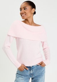 Un pull rose clair à épaules dénudées avec une texture côtelée au niveau de l'encolure et des poignets, fabriqué dans un tissu doux. Associé à un jean en denim bleu.
