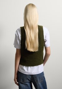 Groene gebreide vest met een cropped ontwerp, gecombineerd met een wit T-shirt. Lange, steile blonde haren vallen omlaag. Jeans zijn zichtbaar daaronder.