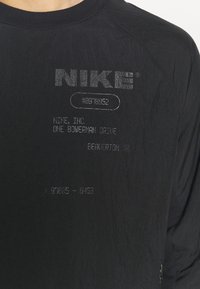 T-shirt noir à manches longues avec logo "NIKE" en relief et texte imprimé, fabriqué dans un tissu texturé et avec un col côtelé. Détail zippé sur le côté.