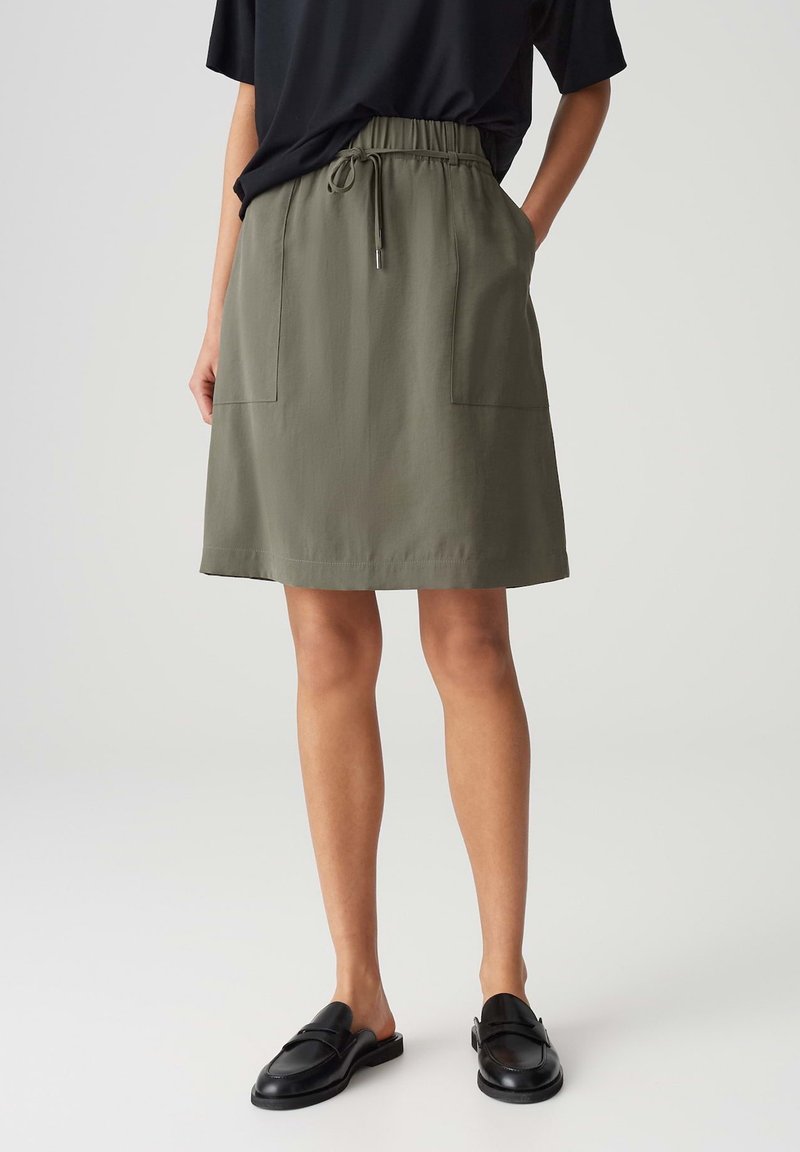 Woman wearing an olive green knee-length skirt with drawstring waist, front pockets, black top, and black slip-on loafers.