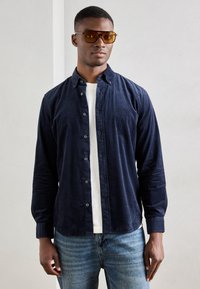 Marineblå corduroy skjorte med knapper og lange ærmer, båret over en hvid t-shirt; parret med lyse denim jeans og brune solbriller.