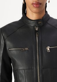 Giubbotto di pelle nera con collo alto, dotato di zip anteriori, due tasche sul petto e dettagli metallici per la chiusura e il design.