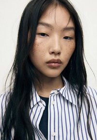 Portrait of a person with long dark hair, wearing a blue and white striped button-up shirt. Noticeable freckles are present on the face.