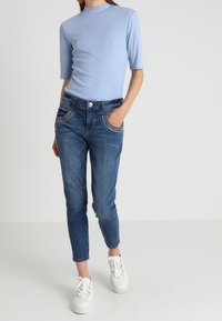 Kvinna som bär en ljusblå kortärmad topp, mörkblå jeans med dekorativa fickor och vita snörskor, står med en hand i fickan.