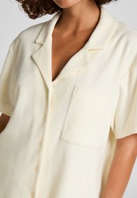 Light cream short-sleeve shirt made from soft fabric, featuring a notched collar, front buttons, and a single pocket on the left chest.