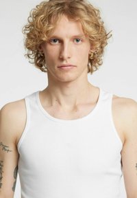 Biały bawełniany top na ramiączkach o prążkowanej fakturze, szerokich ramiączkach i okrągłym dekolcie. Modelka ma kręcone włosy i tatuaże.