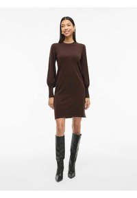 Robe en tricot marron à manches longues bouffantes, avec des poignets côtelés et un col rond, associée à de hautes bottes noires à talons aiguilles.