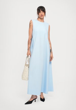 Woman with tattoos wearing sleeveless light blue maxi dress, black heels, and carrying white quilted handbag with gold chain straps.