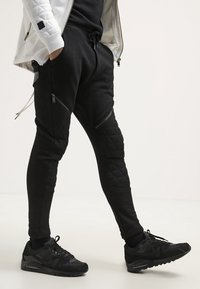 Černé joggingové kalhoty s prošívanými panely, slim fit, a kapsami na zip; doplněné černými sportovními teniskami s texturovanou podrážkou.