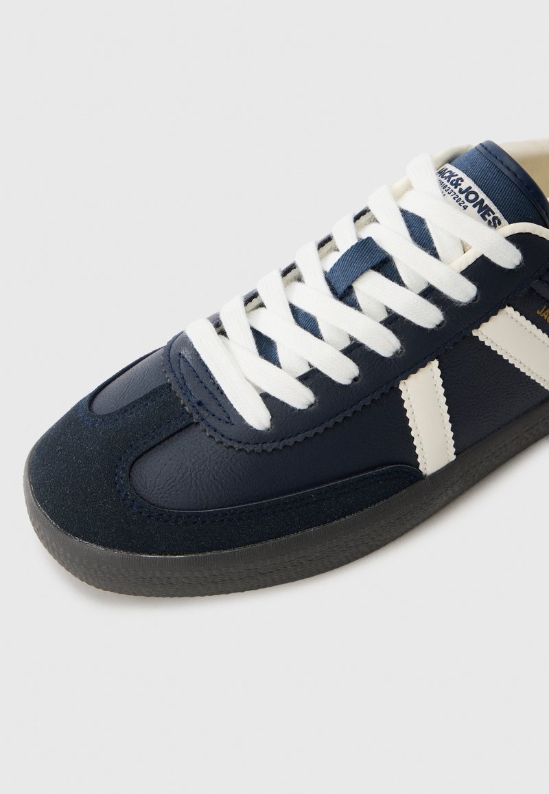 Zapatilla Jack & Jones en navy y blanco con cordones blancos, suela texturizada y rayas laterales en contraste sobre un fondo claro.