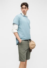 Jeune homme portant une chemise à manches courtes bleu clair sur une chemise à manches longues blanche, un short vert olive, tenant une casquette beige et marron.