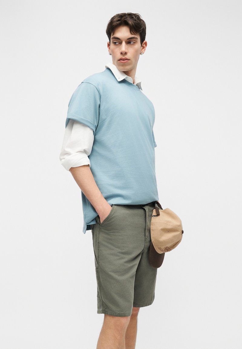 Jeune homme portant une chemise à manches courtes bleu clair sur une chemise à manches longues blanche, un short vert olive, tenant une casquette beige et marron.
