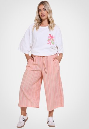Donna bionda in piedi con le mani nelle tasche, indossa una maglietta bianca oversize con grafica rosa, pantaloni larghi a righe rosa e bianchi e scarpe da ginnastica bianche.