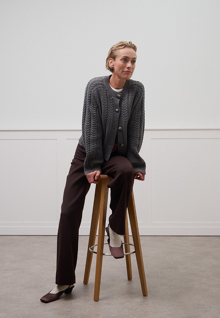 Un cardigan en tricot gris avec des boutons en bois proéminents, associé à un pantalon marron foncé et des chaussures bordeaux. Modèle assis sur un tabouret en bois.