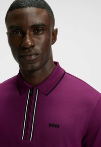 Lila polo t-shirt med dragkedja på kragen, med svarta och vita randiga detaljer och en 'BOSS'-logotyp på vänster sida. Slät textur.