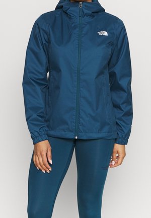 Osoba ubrana w niebieską kurtkę z kapturem North Face na suwak oraz dopasowane niebieskie legginsy, stojąca na jednolitym tle.