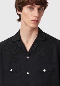 Sort skjorte med blomsterbroderi på kraven, der har to forlommer og firkantede metalaccenter på knapperne. Glat stoftekstur.