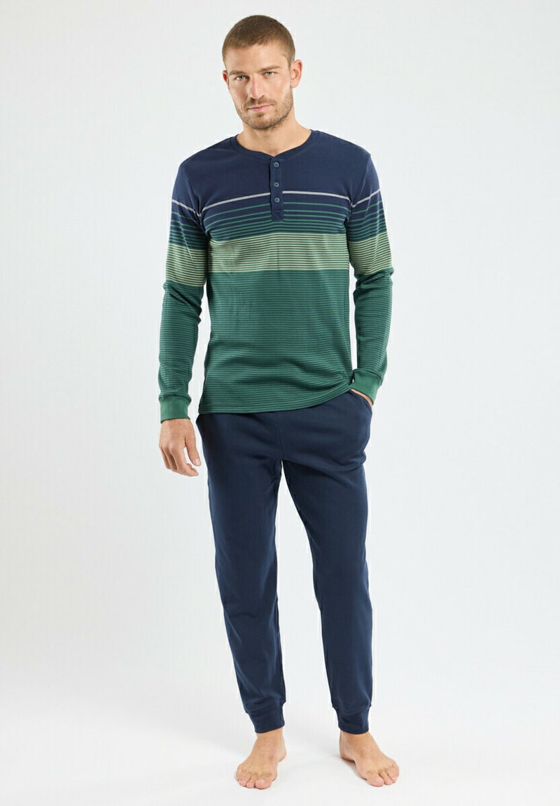 Homme debout pieds nus portant une chemise henley à manches longues rayée verte et marine et un pantalon de jogging marine, sur fond blanc.