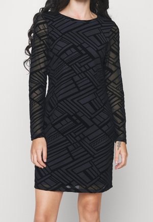 Žena v černých přiléhavých šatech s dlouhými průsvitnými rukávy, zdobenými geometrickým vzorem protínajících se čar a tvarů.