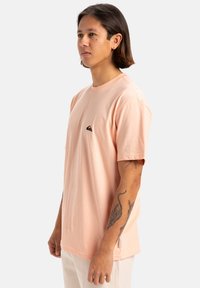 Camiseta de manga corta rosa claro, hecha de algodón, con un pequeño logotipo negro en el pecho, de corte relajado y textura suave.